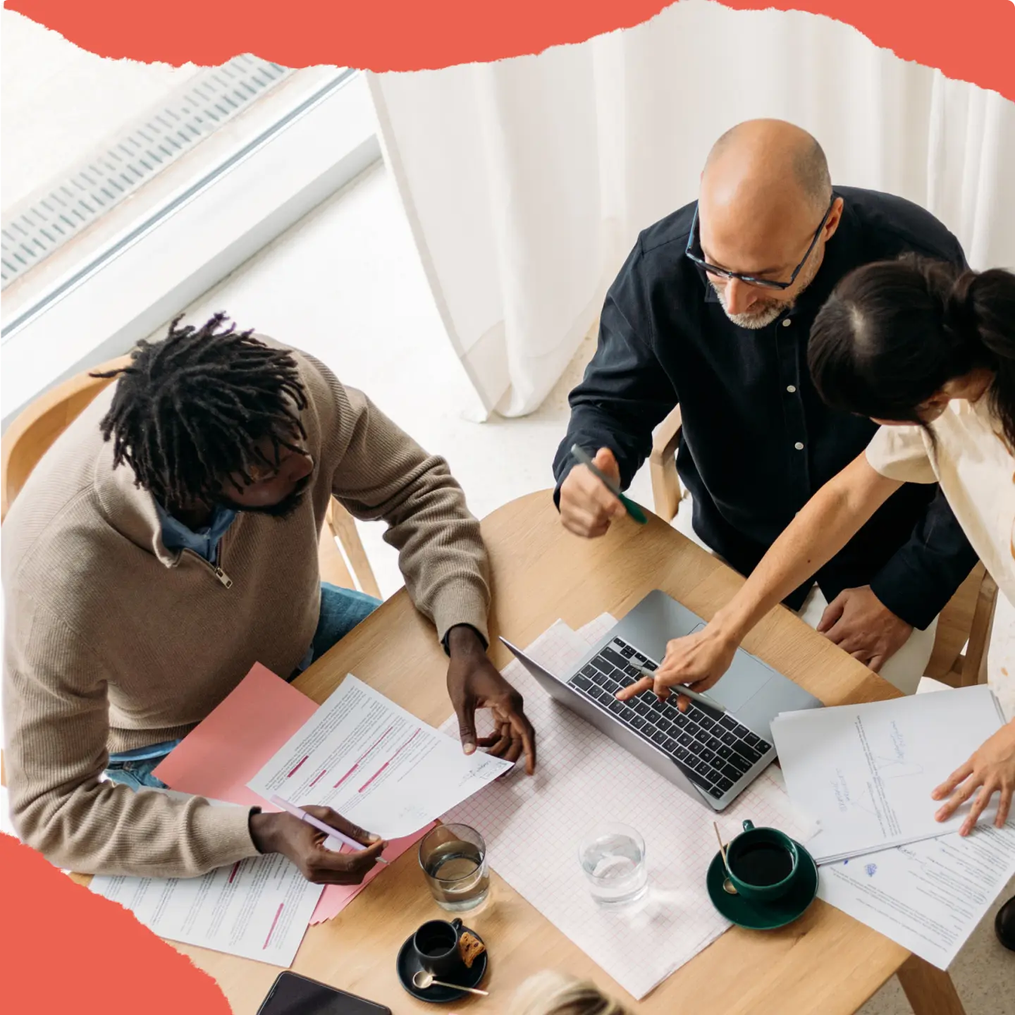 Three people collaborating at a table with documents and a laptop. One person points at the laptop screen.