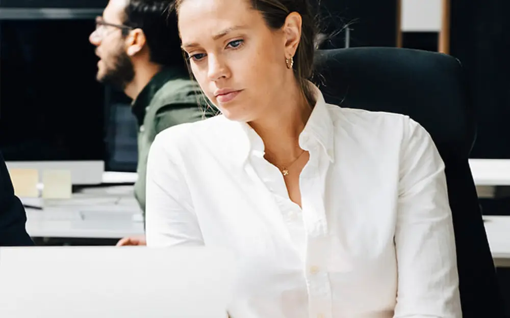 Donna concentrata al computer in ufficio, con un uomo sullo sfondo.
