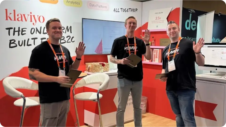 Three people in Klaviyo shirts wave at a booth with Klaviyo branding at an event.