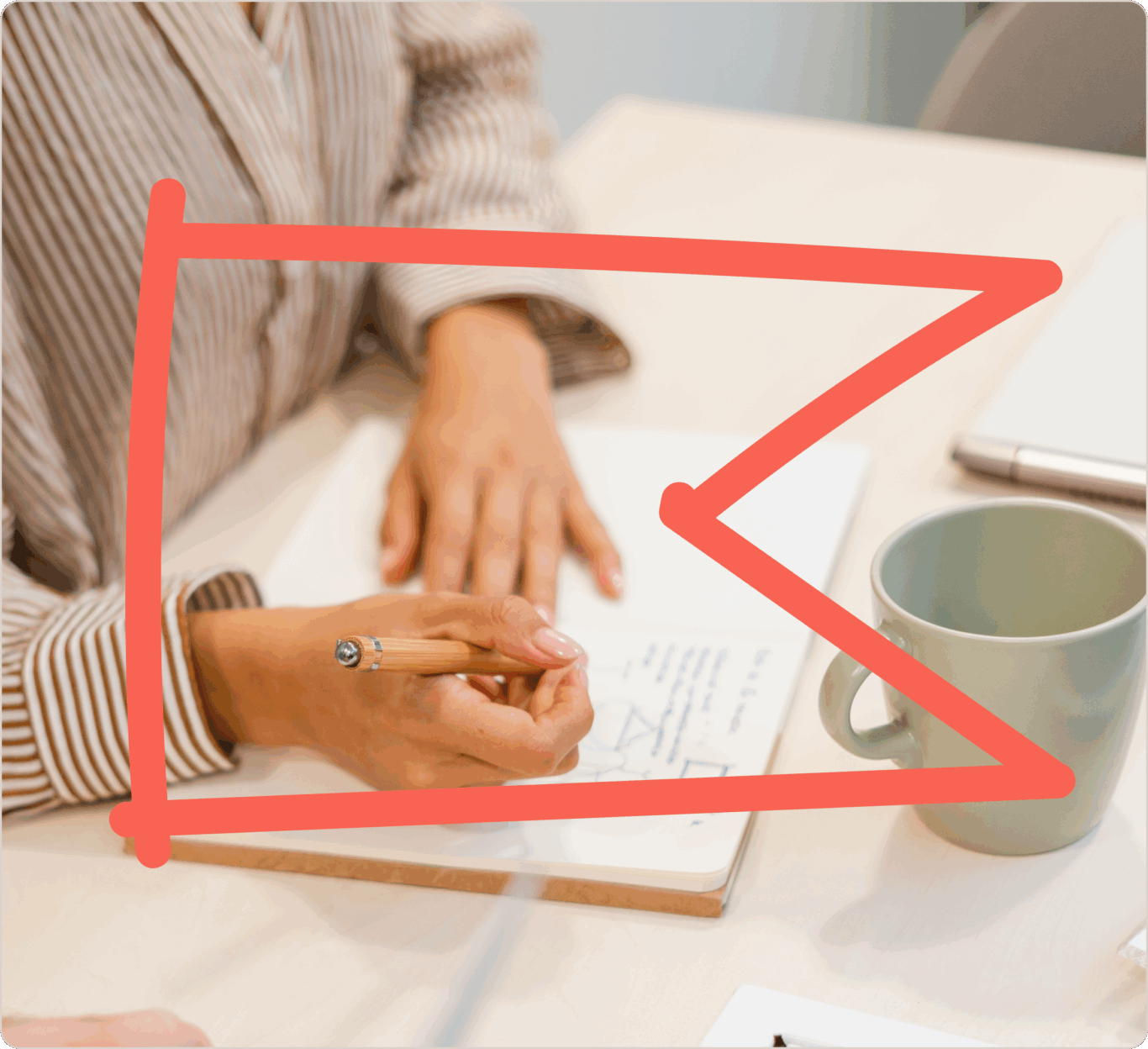 Person writing in a notebook with a pencil, next to a green mug on a table. Red arrow overlay pointing to the notebook.
