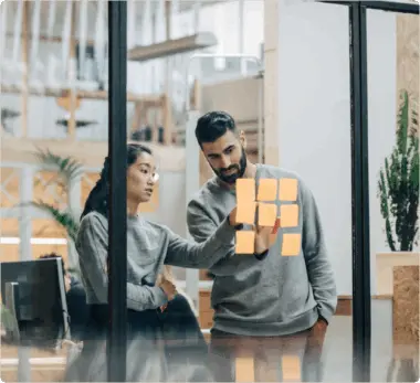 Two people discussing notes on a glass wall in an office setting.