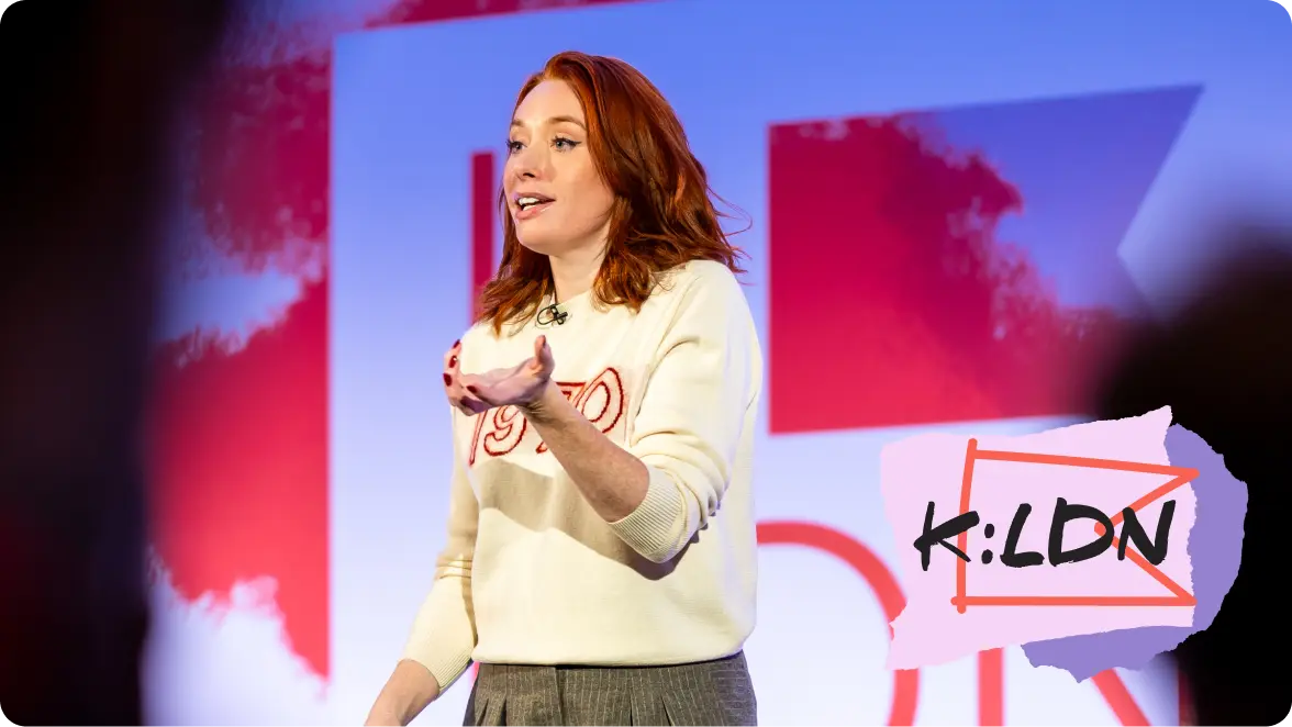 Woman with red hair speaks on stage in front of a red and white backdrop, wearing a cream sweater. The K:LDN logo appears in the bottom right corner.