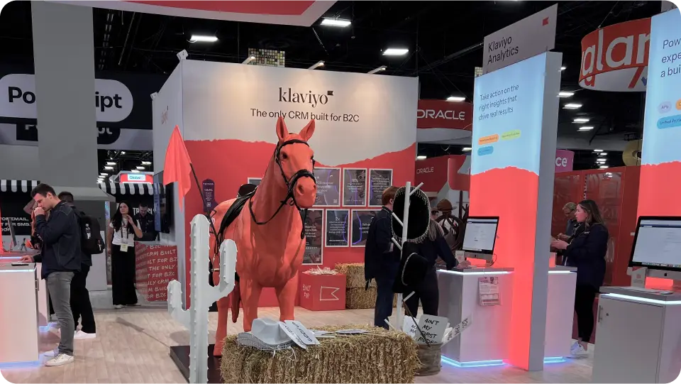 Trade show booth for Klaviyo featuring a bright orange horse statue, hay bales, and signage reading "The only CRM built for B2C." Attendees interact with demo stations and displays promoting Klaviyo Analytics and product features.
