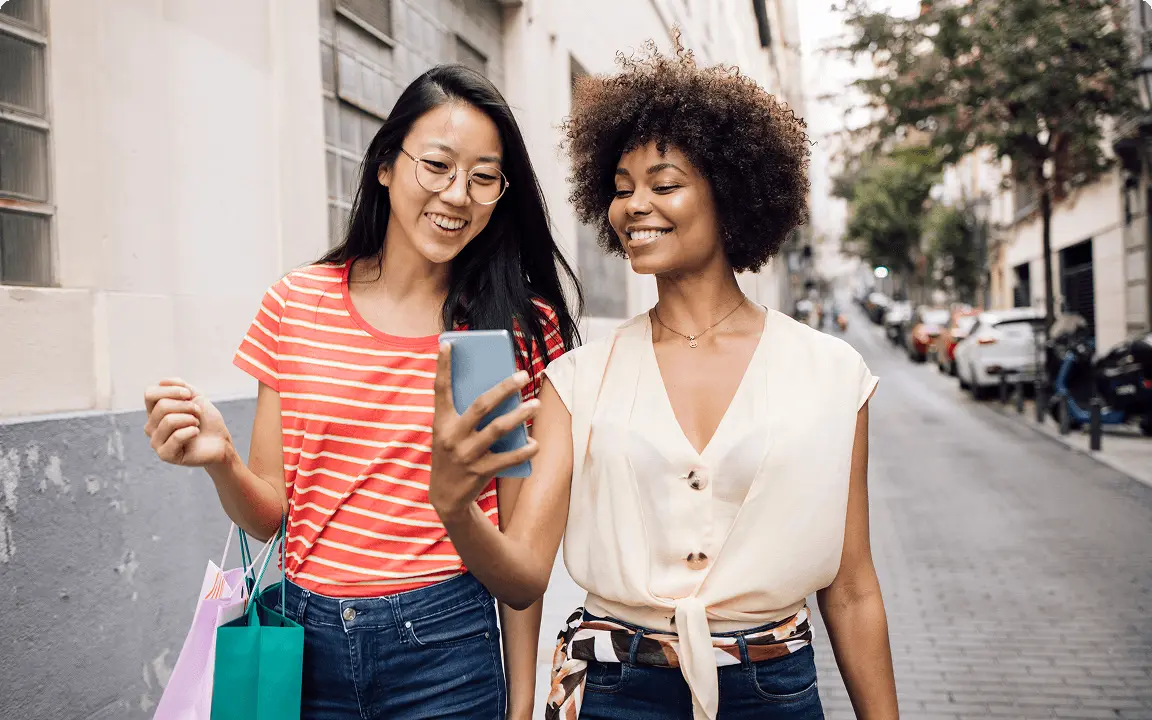 Two women walk down a city street smiling, one holding shopping bags and the other looking at a phone as they chat.