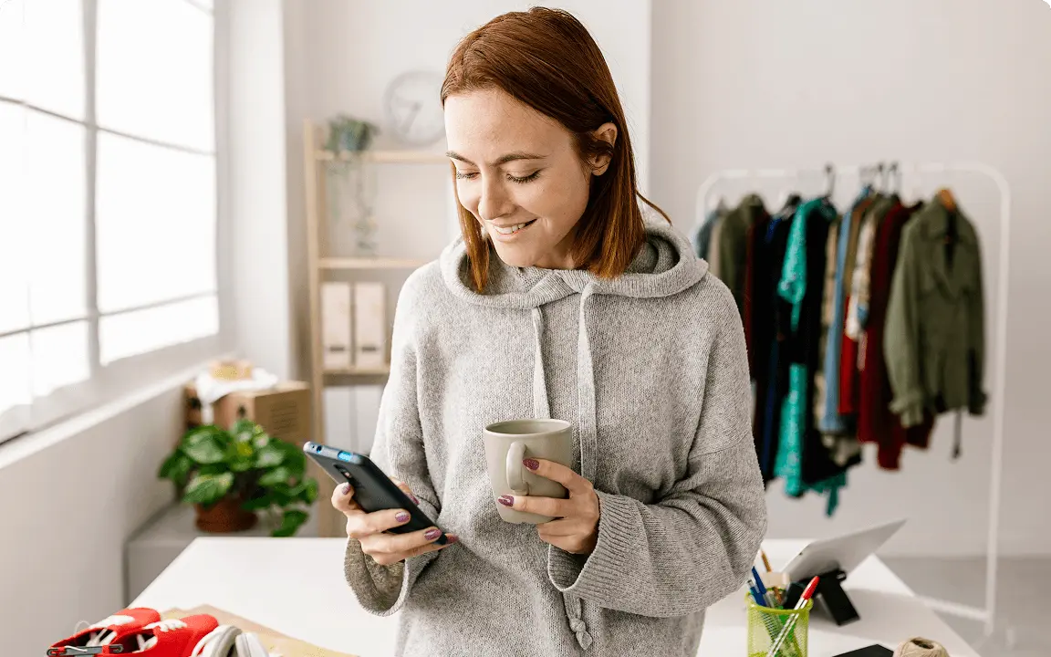 A woman stands in a bright room, smiling while looking at her phone and holding a mug. Behind her is a clothing rack and a desk with various items.