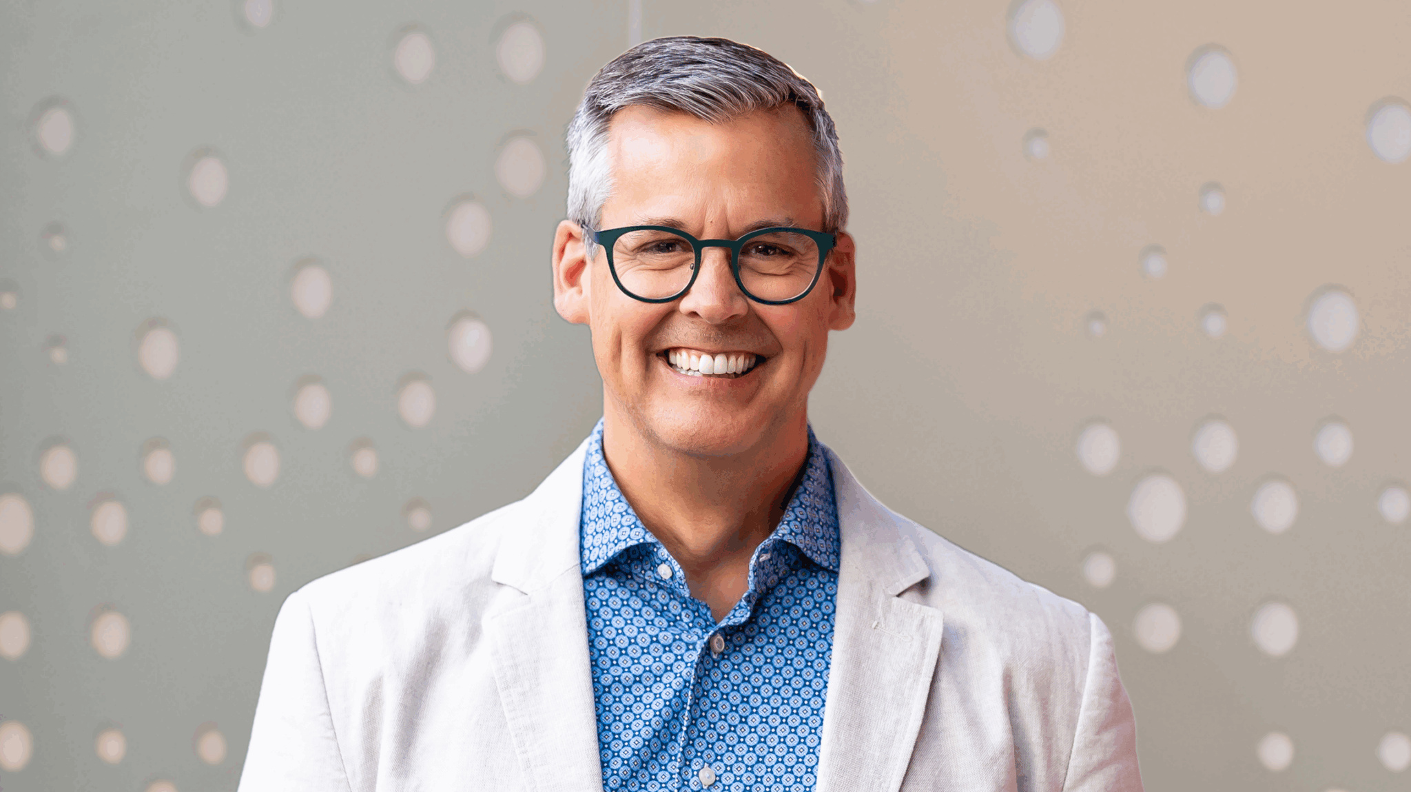 Smiling man with glasses, wearing a light blazer and patterned shirt, standing in front of a dotted wall.