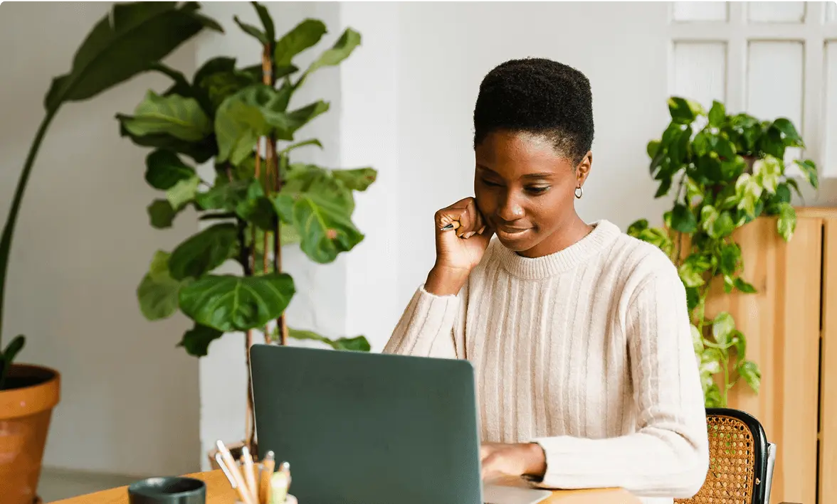 A person in a beige sweater works on a laptop at a desk, surrounded by potted plants.