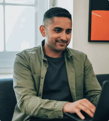 A person in a green shirt is smiling while using a laptop indoors.