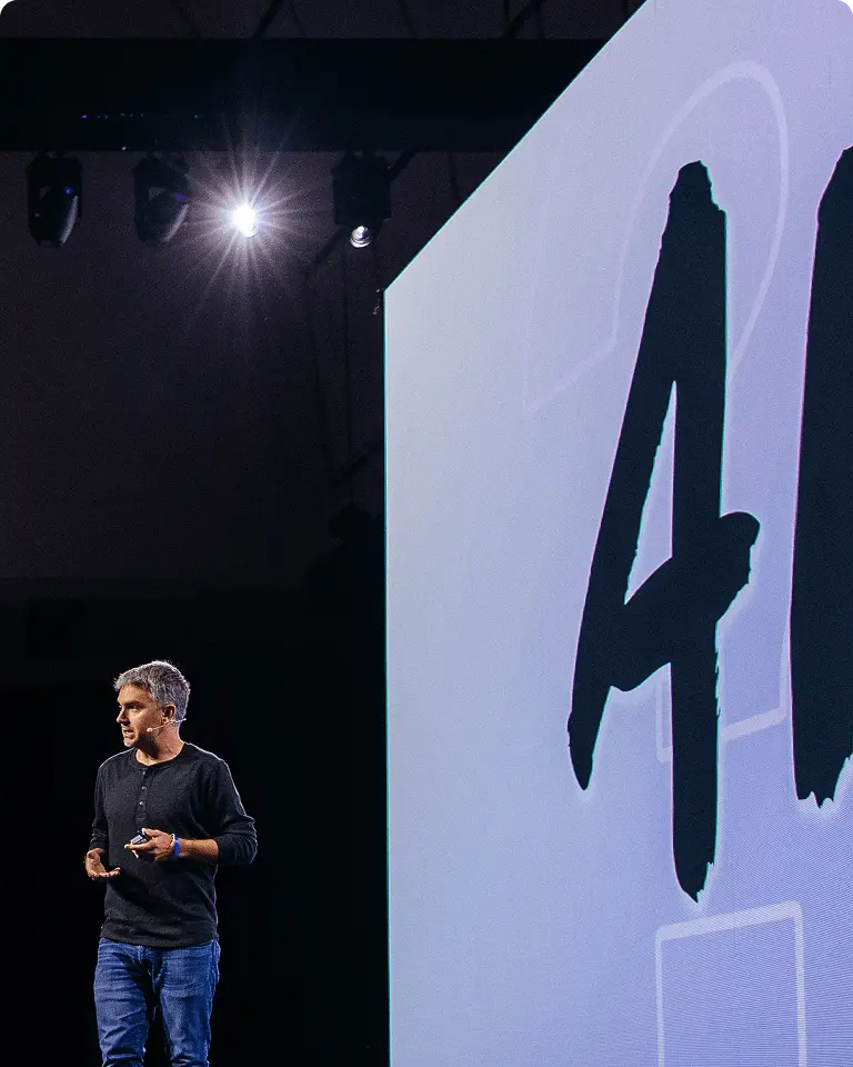 A person speaking on stage next to a large screen displaying the letters "AI".